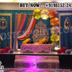 “Vibrant Punjabi Theme Sangeet Stage with colorful umbrellas, traditional Punjabi male and female statues, carved wedding sofa, sequin backdrop, and festive decor by DST Exporters.”