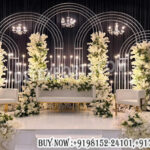 engagement stage decoration, wedding reception stage backdrop, metal arch wedding backdrop, modern wedding stage setup, white floral wedding stage decor, luxury wedding backdrop design, multi-layer arch backdrop wedding, elegant wedding stage panels, candle light wedding stage decor, chandelier wedding stage setup, minimalist wedding stage design, premium event stage backdrop, banquet hall wedding stage decor, wedding decor exporters India, designer wedding stage furniture, floral arch wedding stage, contemporary wedding backdrop panels, luxury engagement stage setup
