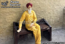 Punjabi Heritage Theme Baba Statue For Selfie Point Realistic Punjabi heritage baba statue wearing yellow traditional attire and red turban, seated on a rustic wooden bench for selfie point and cultural theme decoration.