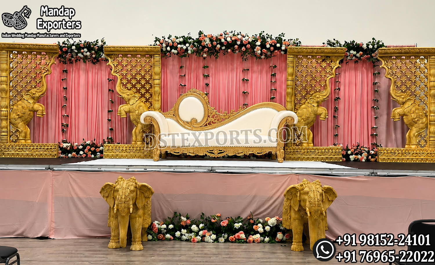 Elegant Indian wedding stage setup featuring a royal white and gold bridal sofa, ornate elephant carved panels, pink drapes, floral arrangements, and decorative elephant statues for traditional ceremonies.